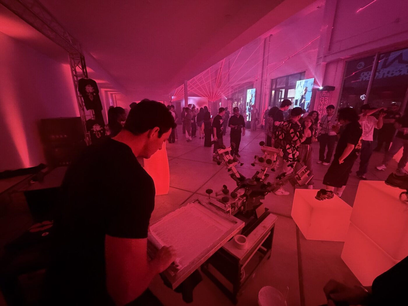 A live screen-printing station running at a packed nighttime brand activation, a printer at the manual press while a crowd fills the room