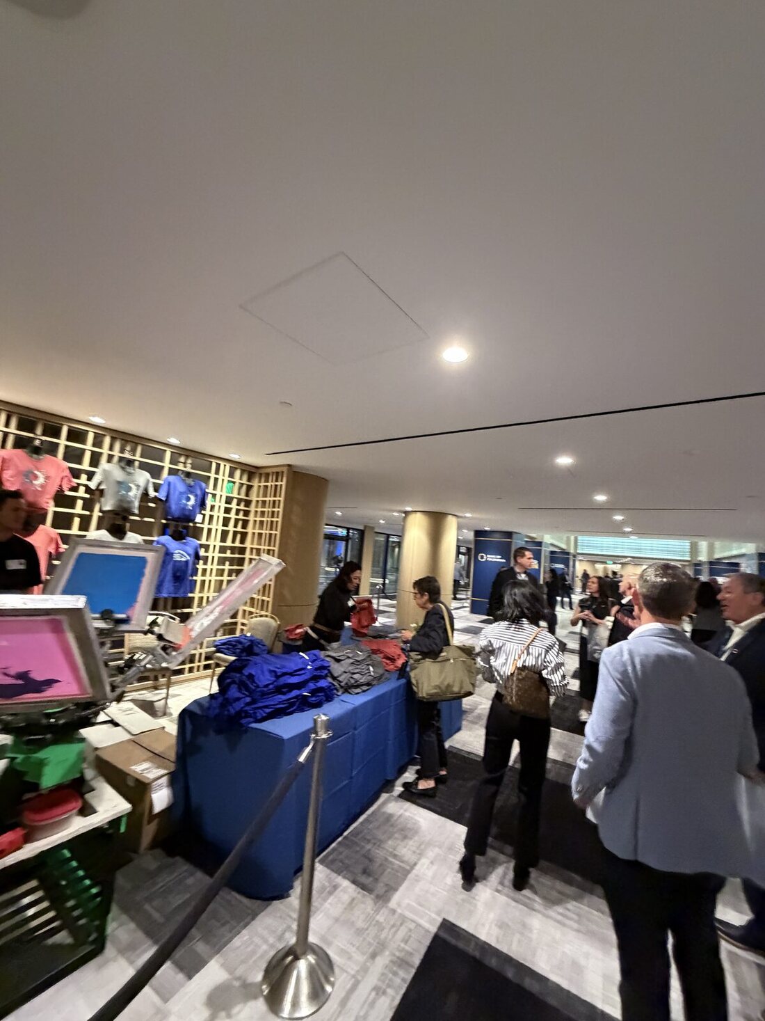 A live-printing booth at a corporate event with a press, a table of folded blank shirts, and printed samples displayed on the wall