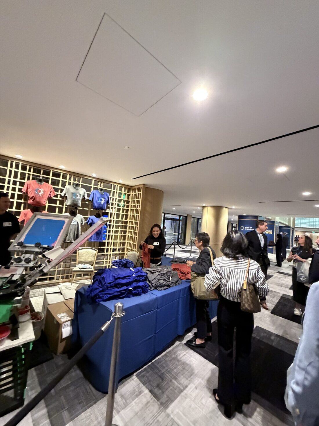 Guests lined up behind a stanchion at a live-printing booth with folded shirts on the table and printed tees on the display wall