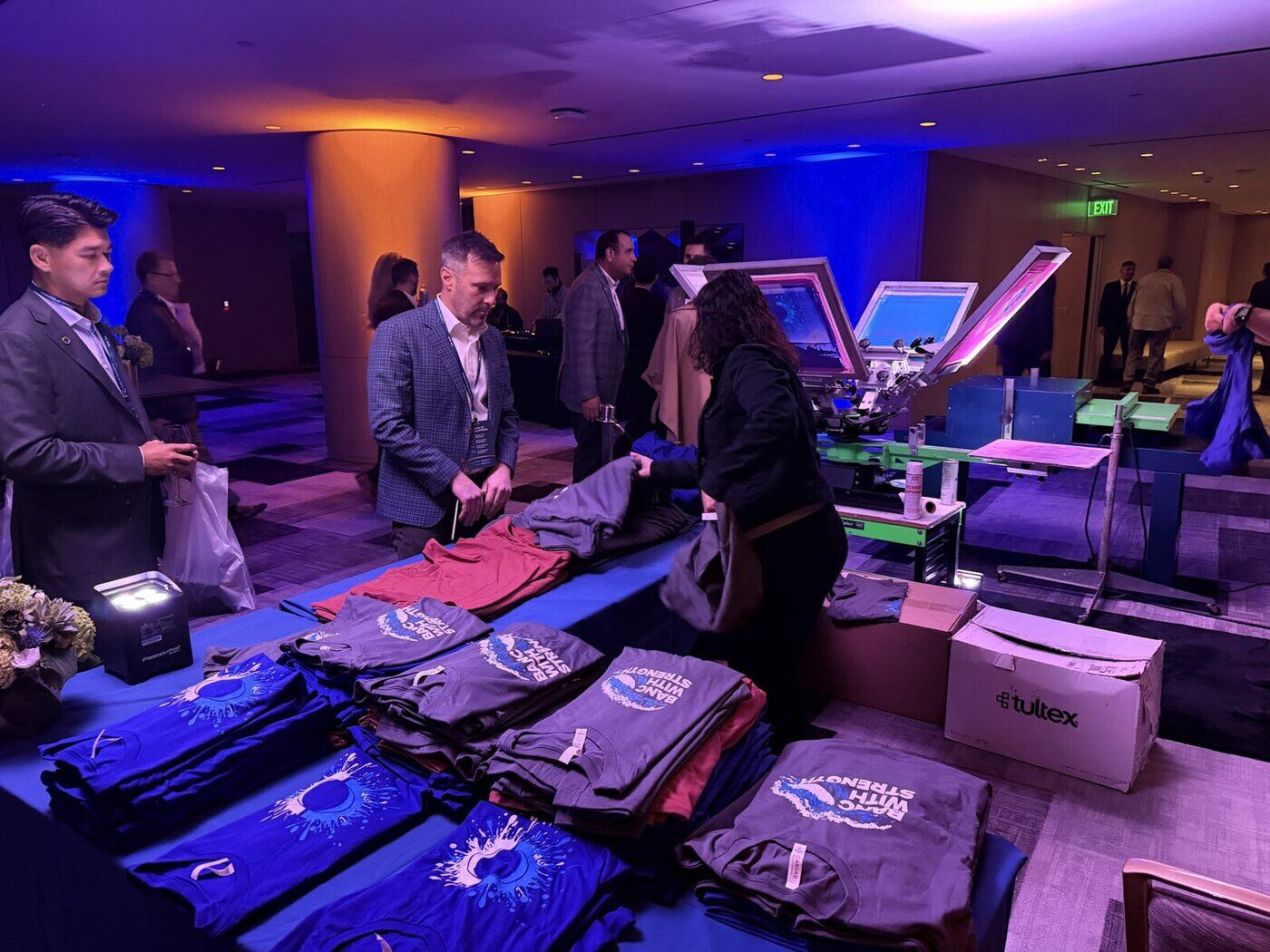 Two screen-printing press stations set up on a Chicago event floor with a table of folded custom event shirts and guests browsing under blue lighting