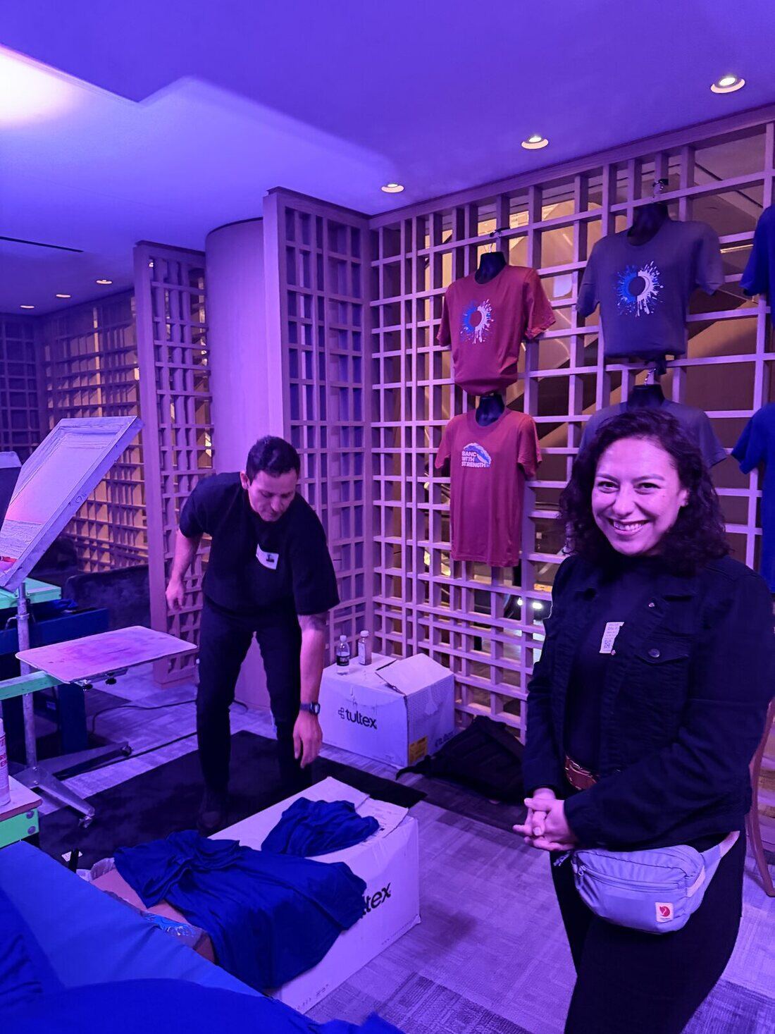 Two Merch Troop crew members setting up a live-printing station, unpacking blank shirts with printed samples displayed on the wall