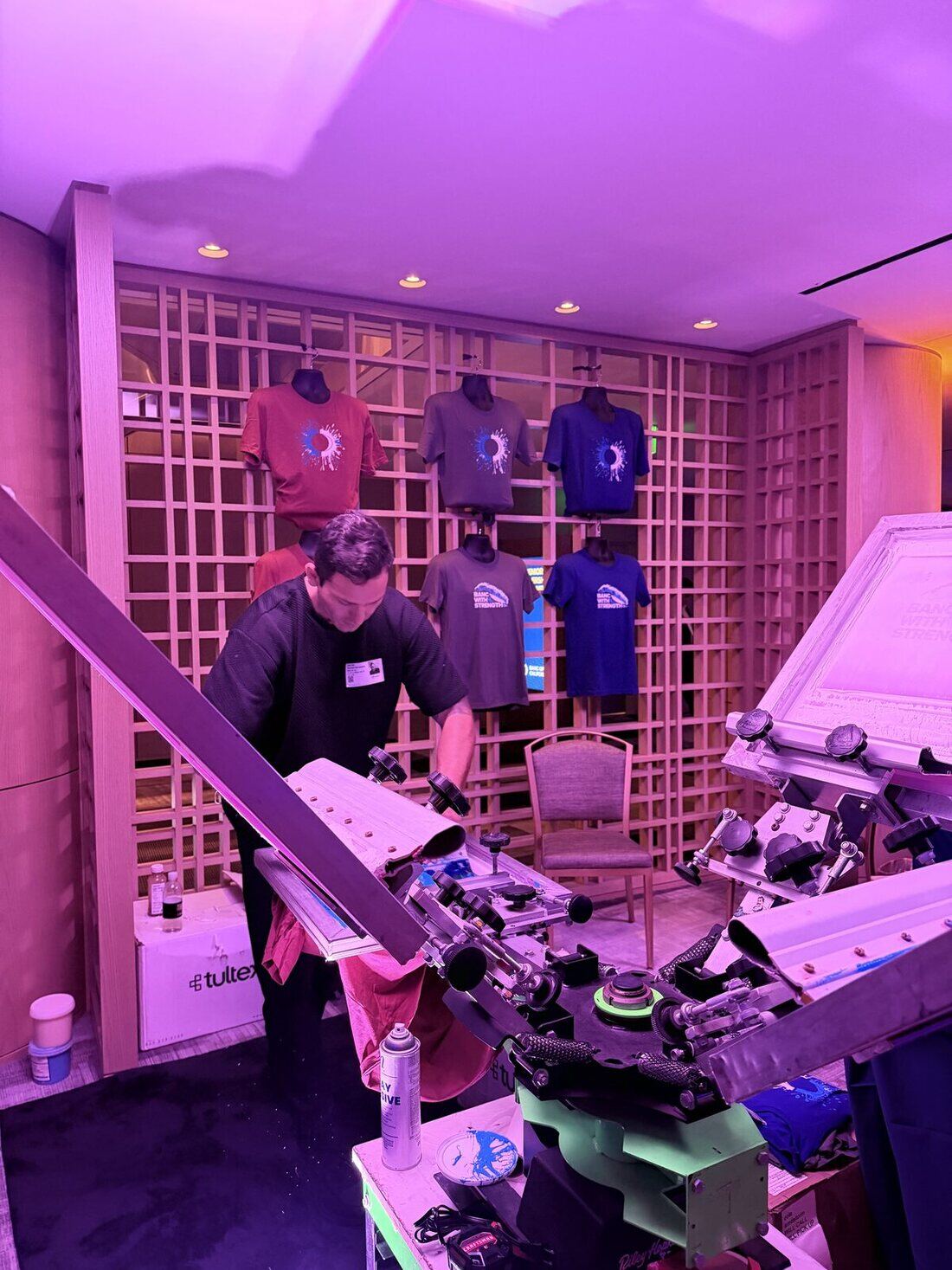 A Merch Troop printer working a manual screen-printing press, with freshly printed event tees hanging on the lattice wall behind