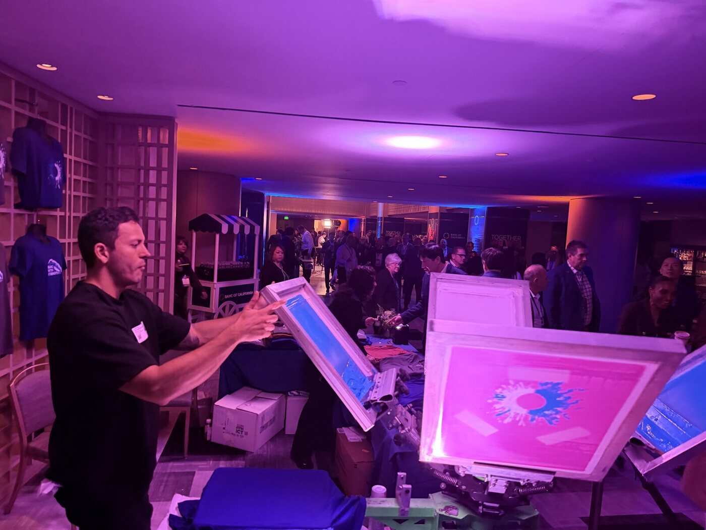 A printer lifting a screen off the press, the printed burst design glowing on the platen, with a crowd gathered at the activation behind